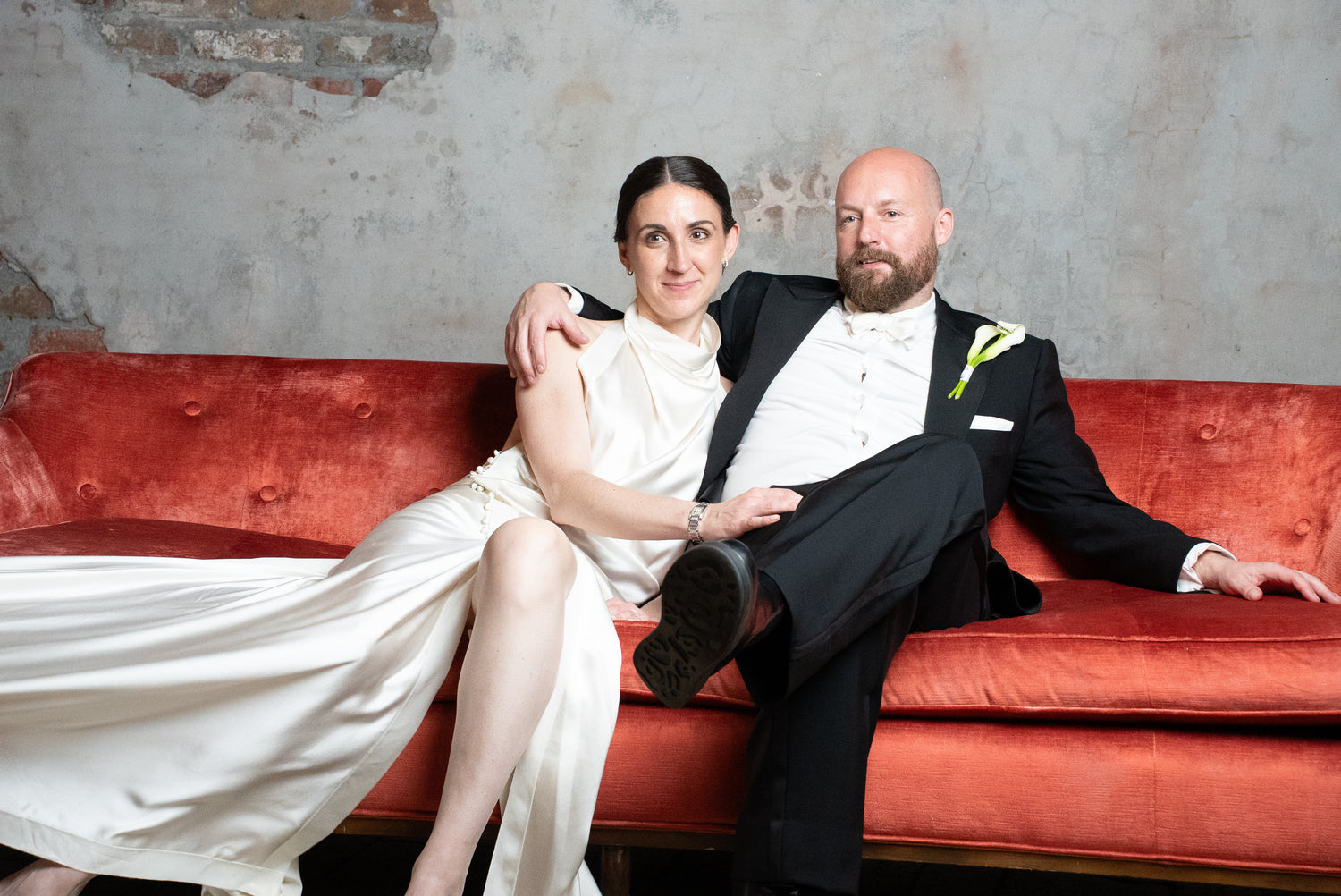 Man in a suit and woman in a white dress sitting on a red couch against a textured wall.